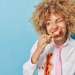 Horizontal shot of curly haired woman bites nugget smeared with ketchup dressed in formal shirt with